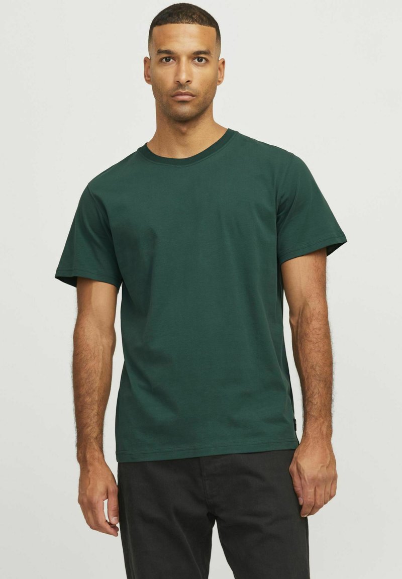 Uomo con capelli corti che indossa una semplice maglietta a maniche corte verde scuro e pantaloni neri, in piedi contro uno sfondo chiaro.