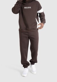 Sweat à capuche marron et jogging assorti avec des accents blancs, arborant un logo sur la poitrine. Tissu doux et coupe décontractée avec des poignets élastiques.