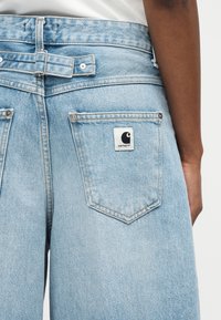 Κοντινό πλάνο παντελονιού από ανοιχτό μπλε τζιν με λεπτομέρεια αγκράφα στη μέση και λογότυπο Carhartt στο πίσω δεξί τσεπάκι.