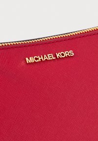Pochette en tissu rouge avec une surface texturée, dotée d'une fermeture éclair dorée et d'un logo "MICHAEL KORS" en évidence.