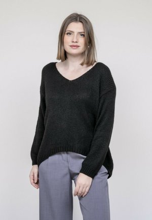Jeune femme aux cheveux mi-longs portant un pull noir à col en V et un pantalon gris clair, se tenant devant un fond clair uni.
