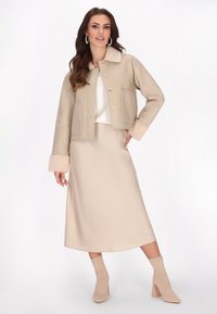 Beige Lederimitat-Cropped-Jacke mit Schafslederkragen und -manschetten, kombiniert mit einem passenden beige Satin-Midikleid und beigen Absatz-Stiefeletten.