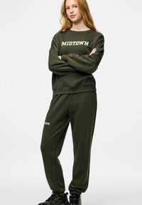 Moteris ilgais plaukais vilki tamsiai žalią džemperį ir sportines kelnes su užrašu „MIDTOWN“, stovi sukryžiavusi rankas, avėdama juodus sportinius batelius.