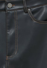 Pantalon en cuir noir avec des coutures contrastantes en marron, une poche avant et un bouton métallique de fermeture à la taille. Texture lisse et design ajusté.