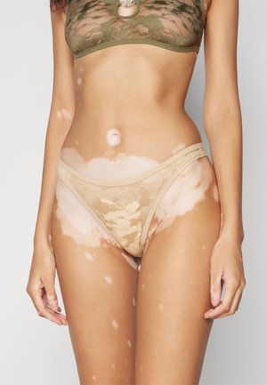 Beige lace thong with floral pattern, featuring a seamless design and delicate side straps, displayed on a model.