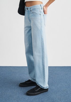 Persoon met lichtblauwe jeans met wijde pijpen, zwarte sneakers, een wit crop top en een zwarte tas op een vloer met blauw tapijt.