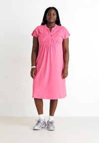 Vestido de algodão rosa com mangas curtas, detalhes em ilhós no corpete, e um corte relaxado que chega até a metade da panturrilha, combinado com sapatilhas prateadas.