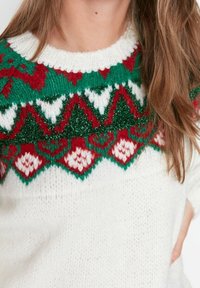 Weißer Strickpullover mit buntem gemustertem Schulterbereich, der rote, grüne und weiße Designs zeigt. Die Textur wirkt weich und dick.