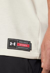 Camisa de algodón color crema con una etiqueta negra que presenta "Under Armour" y "AUTHENTIC" en rojo, adjunta a la costura lateral.