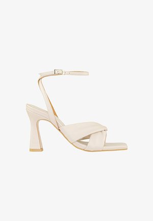 Sandalo alto beige con design a bande incrociate, fascetta alla caviglia, punta aperta e tacco leggermente affusolato con un accento dorato alla base.