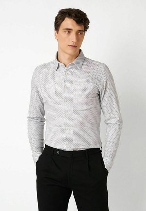 Junger Mann trägt ein weißes gemustertes Hemd mit Knöpfen und schwarze Hose, steht mit den Händen in den Taschen und schaut leicht nach rechts.