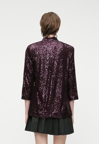 Casaco roxo com lantejoulas, com mangas de três quartos e gola alta, apresentando uma superfície texturizada e brilhante. Usado sobre uma saia preta plissada.