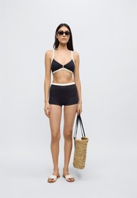 Μαύρο μπικίνι με κρεμ λεπτομέρειες, με τριγωνικό τοπ και σορτς ψηλής μέσης. Πλεκτή ψάθινη τσάντα, φλατ σανδάλια και γυαλιά ηλίου.