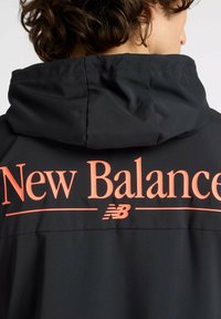 Schwarze Jacke mit Kapuze, die ein auffälliges orangefarbenes "New Balance"-Logo und eine glatte Textur aufweist. Das Design umfasst minimale Hardware-Akzente.