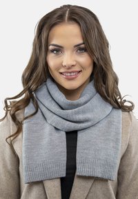 O eșarfă gri deschis dintr-un material moale, cu un bordură ribbed, așezată elegant în jurul gâtului, asortată cu o haină bej.