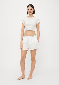 Vit kortärmad crop top med grå textur, tillsammans med vita shorts med ett varumärkestryck på den elastiska midjebandet.