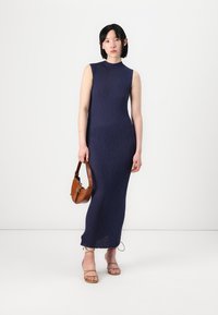 Rochie maxi bleumarin cu o siluetă ajustată, design fără mâneci și fanta laterală. Modelul poartă o geantă maro, completată de sandale bej.
