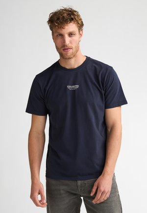 Mann mit lockigem Haar und Bart, der ein dunkelblaues T-Shirt mit weißem Text trägt und vor einem schlichten hellen Hintergrund steht.