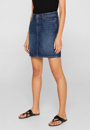 Frau trägt einen dunkelblauen Jeansrock mit Fronttaschen, kombiniert mit schwarzen, flachen Sandalen mit Riemchen und einem schwarzen Oberteil, stehend vor einem grauen Hintergrund.