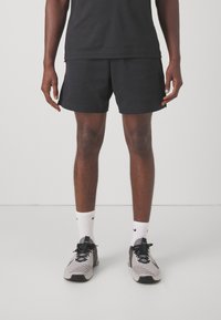 Schwarze Sportshorts mit glatter Textur, kombiniert mit weißen Socken und hellgrauen Turnschuhen mit schwarzen Akzenten und strukturierter Oberfläche.
