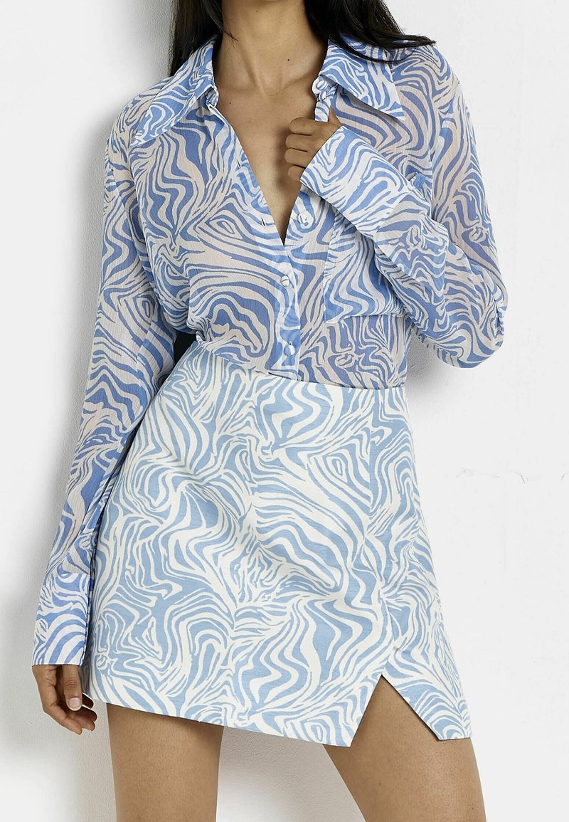 Femme portant une chemise à boutons à rayures zèbre bleu et blanc et une mini-jupe assortie avec une fente à l'avant, posant devant un fond blanc.