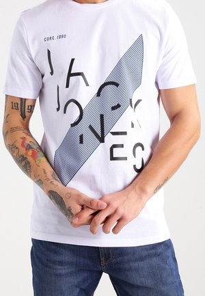 Hvid kortærmet t-shirt med sort geometrisk tekst og et diagonal stribet mønster. Båret med blå jeans, vist på en stående model.