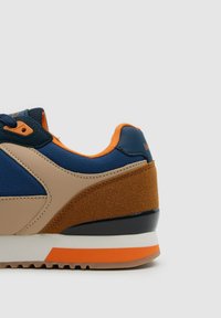 Vista lateral de una zapatilla deportiva azul, beige y marrón con forro y detalles naranjas en la suela, sobre un fondo gris claro liso.