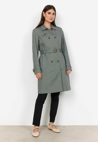 Frau in einem grauen Trenchcoat mit Gürtel, schwarzen Hosen und beigen Ballerinas vor einem schlichten weißen Hintergrund.