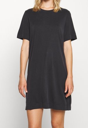 Femme portant une robe T-shirt noire à manches courtes et ample, debout devant un fond clair uni.