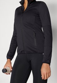 Chaqueta atlética negra con cuello alto, cremallera completa, bolsillos laterales con cremallera y tela elástica. Estilo ajustado, combinada con leggings negros.