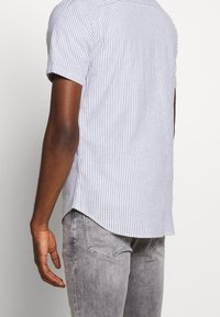 Košile s krátkým rukávem v modro-bílém pruhu, zakřivený lem, materiál směs bavlny, nošena s světle šedými džíny, zobrazeno boční a zadní pohled.