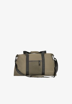 Homme en tenue noire portant un grand sac vert olive avec bandoulière, debout devant un fond clair uni.