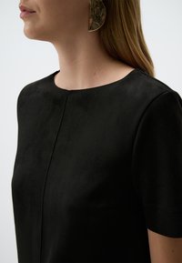 Zwart suede top met korte mouwen, voorzien van een ronde halslijn en een subtiel naaddetail. Gecombineerd met een grote, metalen oorbel.
