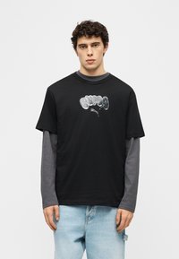 Νέος άνδρας με σγουρά μαλλιά, φορώντας μαύρο T-shirt Puma πάνω από γκρι μπλούζα μακρυμάνικη και ανοιχτόχρωμα γαλάζια τζιν, στέκεται μπροστά σε λευκό φόντο.