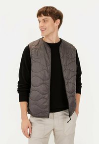 Graue gesteppte Weste mit rundem Ausschnitt und Reißverschluss, über einem schwarzen Pullover getragen. Das Model steht vor einem neutralen Hintergrund.