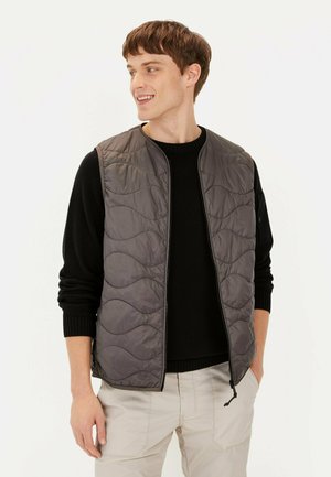 Graue gesteppte Weste mit rundem Ausschnitt und Reißverschluss, über einem schwarzen Pullover getragen. Das Model steht vor einem neutralen Hintergrund.