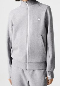 Šedá mikina na zip z měkké tkaniny, s vysokým límcem, bočními kapsami a malým logem na hrudi. Hladká textura.