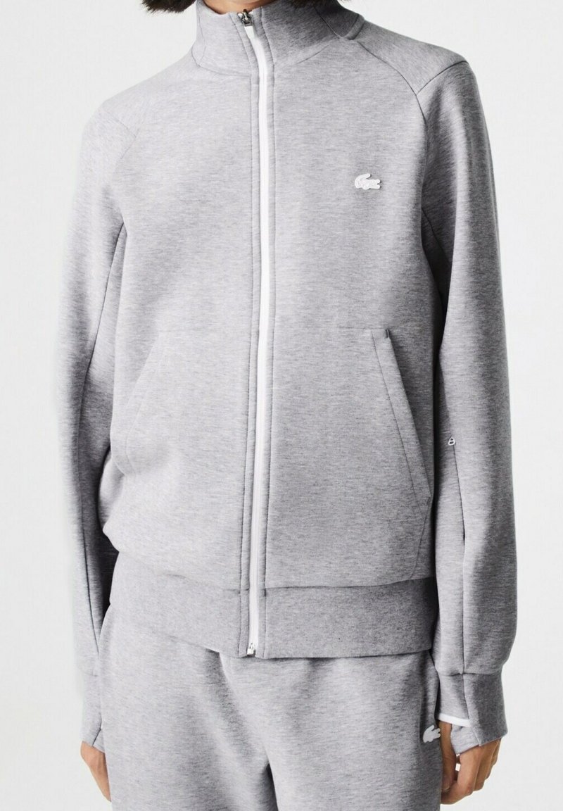 Šedá mikina na zip z měkké tkaniny, s vysokým límcem, bočními kapsami a malým logem na hrudi. Hladká textura.