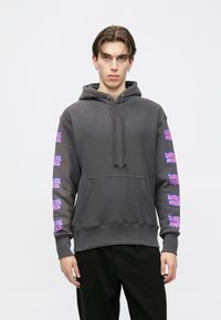 Grauer Kapuzenpullover mit Kordeln und Fronttasche. Der Ärmel zeigt sich mit wiederholtem pinkfarbenem grafischem Text. Der Stoff wirkt weich und strukturiert.