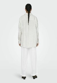 Camicia oversize a righe bianche con yoke sul retro e maniche lunghe, abbinata a pantaloni larghi in lino bianchi e scarpe con tacco nere.