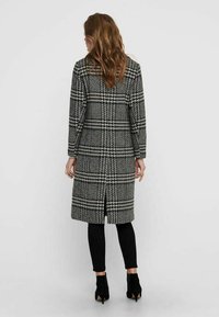 Langer, karierter Mantel mit schwarz-weißem Muster, mit einem Schlitz in der Mitte des Rückens und langen Ärmeln, kombiniert mit engen schwarzen Hosen und Stiefeletten.