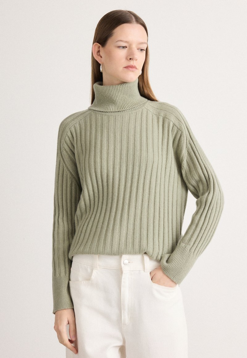 Vihreä ribattua turtleneck-pulloveria, jossa on pitkät hihat ja rento fit, yhdistetty valkoisiin korkea vyötäröisiin housuihin.