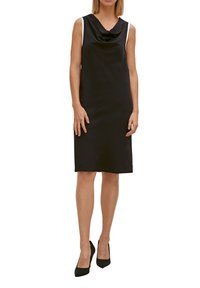 Schwarzes ärmelloses Kleid mit Wasserfall-Ausschnitt und weißen Schulterakzenten; knielang mit einem glatten, strukturierten Stoff. Schwarze High Heels.