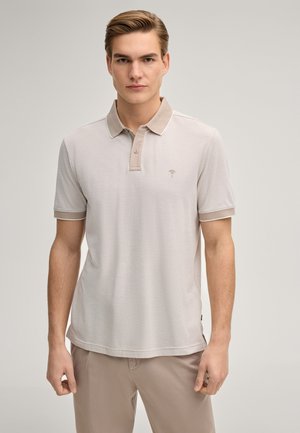 Giovane uomo con capelli castano chiaro, indossa una polo beige e pantaloni coordinati, in piedi davanti a uno sfondo neutro con un'espressione neutra.