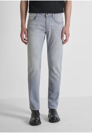 Persona che indossa jeans aderenti grigio chiaro e stivali alla caviglia in pelle nera, in piedi davanti a uno sfondo bianco semplice.