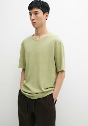 Junger Mann mit kurzen schwarzen Haaren, der ein hellgrünes kurzärmeliges Hemd und dunkelbraune Hosen trägt und vor einem weißen Hintergrund steht.