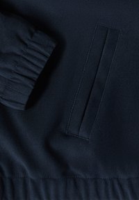Section de veste bleu marine avec un poignet côtelé et une poche latérale, caractérisée par une texture lisse et des détails de couture subtils.