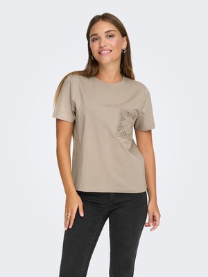 Vrouw draagt een beige T-shirt met korte mouwen en een gehaakt zakje op de linkerborst en zwarte spijkerbroek, staand tegen een effen achtergrond.