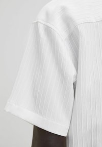 Primo piano di una camicia bianca a maniche corte con texture su un braccio di pelle scura, che mostra un motivo a righe verticali e l'orlo della manica piegato.