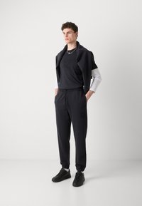 Black cotton crew-neck t-shirt with embossed logo and matching joggers. Sweatshirt draped around shoulders. Sneakers complete the outfit.